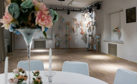 Арендовать Пространство для ваших праздников. Просторное помещение с деревянным полом, белыми стенами, круглым столом с белой скатертью, цветами и свечами. На стенах и стойках декоративные цветочные композиции. Освещение точечное, присутствует оборудование для световых эффектов.