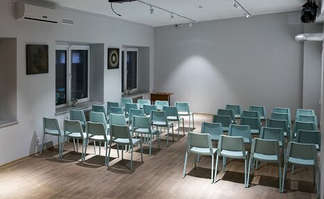 Арендовать Пространство для ваших праздников. Просторная комната с серыми стенами и деревянным полом, расставленные в ряды голубые пластиковые стулья, два окна с подоконниками, потолочный регулированный свет, настенные декоративные элементы, кондиционер, вытяжка, небольшой деревянный стол.