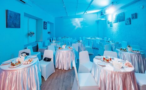 Арендовать Пространство для ваших праздников. Банкетный зал с круглымі столами, покрытыми белыми скатертями, и белыми стульями. На столах блюда и салфетки. Синие стены с картинами. Пол деревянный. Освещение яркое, из потолочных светильников. Настенная декорация из цветов.