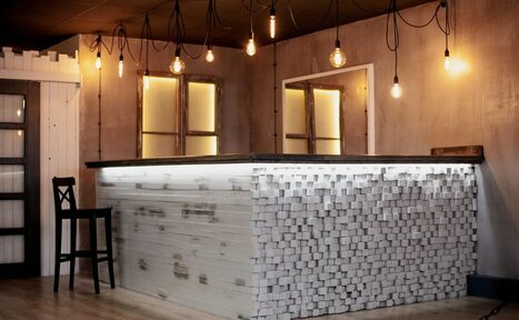 Арендовать Лофт-пространство в Ярославле. Барная стойка с отделкой белыми кубами и черной столешницей, барный стул, деревянные стены с индустриальными светильниками на проводах, два окна с подсветкой, деревянный пол, матовое освещение.