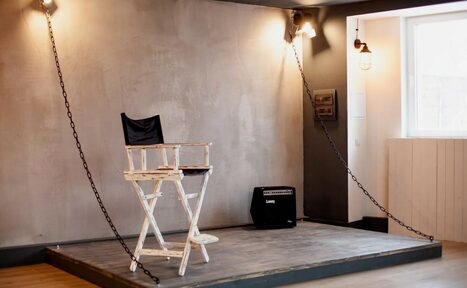 Арендовать Лофт-пространство в Ярославле. Небольшая сцена с высоким белым деревянным стулом режиссера, рядом усилитель. Серые стены, светлое деревянное напольное покрытие. Освещение представлено лампами на потолке и у окна. Натянуты декоративные цепи. Окно с белой рамой справа.