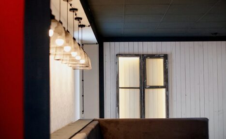 Арендовать Лофт-пространство в Ярославле. Небольшое пространство с белыми деревянными панелями и обшитыми стенами, коричневый диван, ряд висящих лампочек с абажурами, окно без стекол, темный потолок.