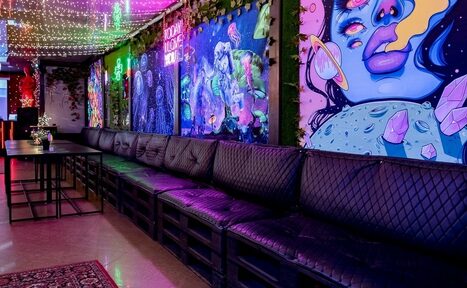 Арендовать Яркий лофт для праздников. Лофт-пространство с длинным деревянным столом и черными кожаными диванами. Яркие граффити на стенах. Подвесные гирлянды разноцветных ламп. Напольное покрытие из светлого ламината. Зеленые декоративные растения вдоль стен.