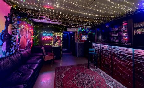 Арендовать Яркий лофт для праздников. Лаунж-пространство с барной стойкой, черной кожаной мебелью и деревянными элементами отделки. Потолок украшен гирляндами. На стене яркие граффити и неоновая вывеска. Ковровое покрытие с орнаментом на полу.