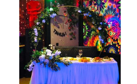 Арендовать Яркий лофт для праздников. Пространство с праздничным декором, длинный стол с белой скатертью и блюдами, арка из зелени и белых цветов. На стене яркий абстрактный узор и постер с надписью "Танцы". Разноцветное освещение, стул с подушкой, блестящая занавеска.