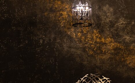 Арендовать Лофт со своим двориком. Темная стена с текстурой и золотистыми акцентами, подвесной светильник в форме клетки с лампой внутри, металлический декоративный элемент на переднем плане. Освещение теплое и направленное.