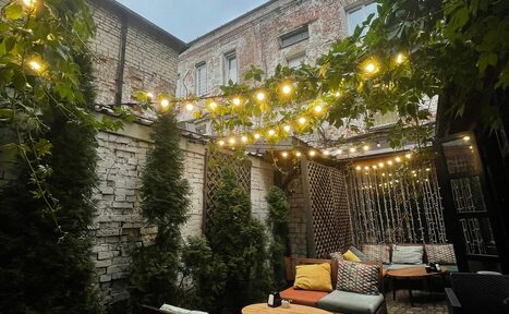 Арендовать Лофт со своим двориком. Уличное пространство с кирпичными стенами, выложенной плиткой площадкой и диванами с подушками. Множество растений и гирлянд с лампочками. Деревянные круглые столы, мягкие сиденья, разноцветные подушки. Вертикальные решетки с растениями.