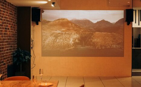 Арендовать Антикафе с несколькими залами. Помещение с кирпичной стеной слева и гладкой стеной, на которой проецируется изображение горного пейзажа. Пол с крупной плиткой. Стол из дерева с двумя бокалами и пустой тарелкой. Потолочные светильники обеспечивают мягкое освещение.