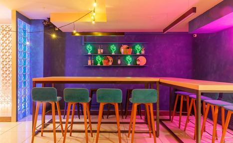 Арендовать Антикафе с несколькими залами. Барное пространство с высоким столом и стульями с зелеными сиденьями. Фиолетовая стена с полкой, на которой расположены неоновые фигуры кактусов и бутылки. Яркое освещение с теплым и фиолетовым оттенком, плиточный пол.