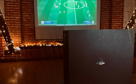 Арендовать Лофт-мансарда . Кирпичная комната с деревянным полом, на стене проектор показывает футбольный матч. В центре перед проектором черная игровая консоль. По периметру стены и вокруг экрана развешена гирлянда с огнями.