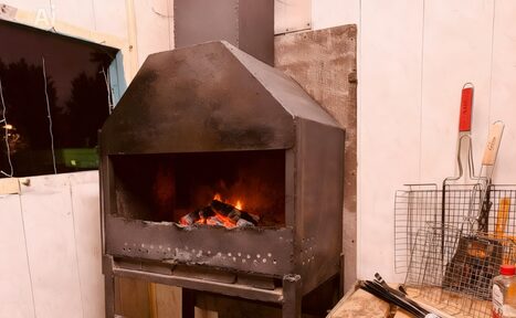 Арендовать Лофт-мансарда . Камера с металлическим камином или мангалом, в углу великая металлическая решетка и инструменты для барбекю. Стены выкрашены в светлый цвет, у окна видна тёмная занавеска. Справа на деревянной поверхности размещена ёмкость.