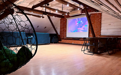 Арендовать Лофт-мансарда . Чердачное помещение с наклонным потолком. Пол из светлого дерева. Красные кирпичные стены. Синий диван, подвесное кресло, стол с черными стульями. На стене экран с проектором. Потолочные деревянные балки. Светильники на потолке.