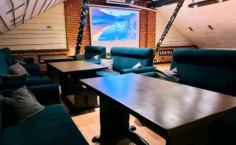 Арендовать Лофт-мансарда . Мансарда с кирпичными стенами и деревянными панелями, с синими мягкими диванами, рядом несколько прямоугольных столов. Большой экран проектора на фоне. Потолок с черными балками. Теплое освещение, гирлянды на стойках. Пол светлый деревянный.