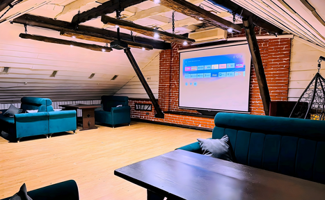 Арендовать Лофт-мансарда . Чердачное пространство с деревянным полом и открытыми балками, кирпичная стена с большим экраном, синие диваны и кресла, черный стол, плетеное кресло-качалка, яркое точечное освещение, белые и серые панели на стенах.