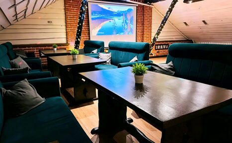 Арендовать Лофт-мансарда . Кофейня с деревянными панелями и кирпичной стеной. Темные диваны и столы. На столах зеленые растения в горшках. На стене проектор с изображением природы. Потолок с косыми линиями обрамлен гирляндами. Светлый деревянный пол.