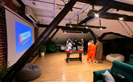 Арендовать Krisha loft (Крыша Лофт). Лофт-пространство с деревянными полами и кирпичной стеной. На стене проекционный экран, на полу пуфы и столик с красными стаканами. Пучковая лампа, деревянные балки, зеленый диван с подушками, группа людей.