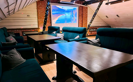 Арендовать Лофт-мансарда . Лофт-пространство с деревянными столами и темными диванами. Красный кирпич и деревянные панели на стенах. Проектор с изображением на экране. Деревянный пол, обшитый потолок. Ограниченное освещение с гирляндами. Несколько подушек на диванах.