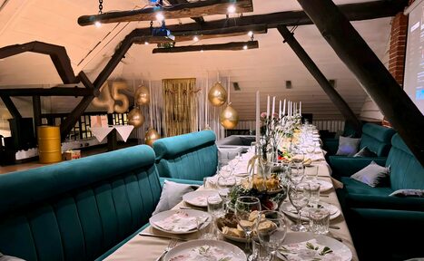 Арендовать Krisha loft (Крыша Лофт). Чердак с деревянными балками, длинный стол с белыми скатертями, сервировка тарелками и бокалами. С каждой стороны стола зелёные диваны, декоративные золотые шары, свечи, проектор, светлая стена, элементы кирпичной кладки.