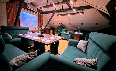 Арендовать Krisha loft (Крыша Лофт). Лофт-пространство с деревянным потолком и балками, кирпичная стена. Мягкие зелёные диваны вокруг деревянного стола с сервировкой. Проектор на экране. Подвесной светильник. Плетёное кресло в углу. Пол светлый, деревянный.