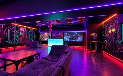Арендовать Cyberpunk (Киберпанк). Лофт с неоновым освещением. Г-образный диван, журнальный столик, барная стойка с табуретами. Пол тёмного дерева. На стенах граффити в стиле киберпанк. Потолок декорирован лампами и металлическими конструкциями.