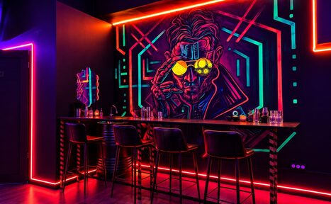 Арендовать Cyberpunk (Киберпанк). Коктейльный бар с неоновым освещением. Высокий барный стол и пять стульев. На стене неоновое граффити с изображением человека в очках. Пол тёмного цвета, контурное освещение вдоль стен. На барной стойке расставлены бокалы и бутылки.
