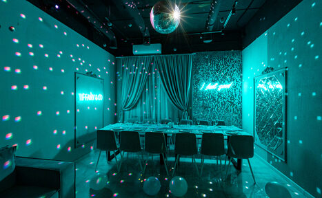 Арендовать Тиффани. Изысканный лофт с мебелью, светомузыкой и караоке. Пространство для мероприятий с длинным столом и стульями. Светодиодное освещение, неоновая надпись "TIFFANY & CO", зеркала, светло-бирюзовые шторы, диско-шар. Потолок чёрный с открытыми коммуникациями. На полу белые шары.