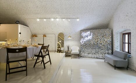 Арендовать Brodniki Loft (Бродники Лофт). Лофт из белого кирпича. Арендуемое пространство с белыми кирпичными стенами и деревянным полом. Зона с накрытыми столами и стульями, светло-серый диван и зеркало. Сверху точечные светильники. На стене светящаяся надпись "Happy Birthday".