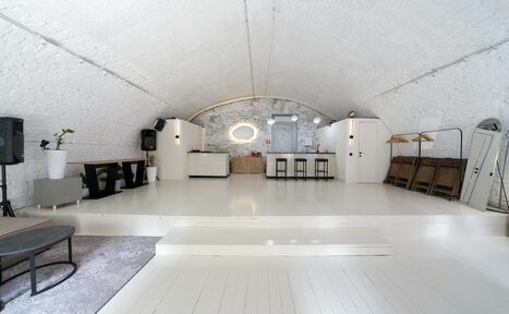 Арендовать Brodniki Loft (Бродники Лофт). Лофт из белого кирпича. Арочное пространство с белым кирпичным сводом. В центре сцена с барной стойкой и стульями. Черно-белая мебель, зеркала и аудиосистема. Деревянный пол и вешалки справа. Светлая цветовая гамма, хорошее освещение.