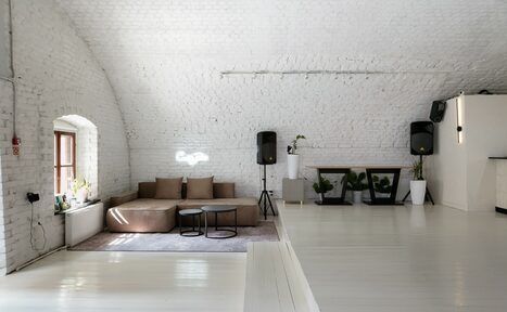 Арендовать Brodniki Loft (Бродники Лофт). Лофт из белого кирпича. Лофт-пространство с белыми кирпичными стенами и арочными потолками. Есть диван с подушками на ковре, два маленьких столика, растительность в белых вазах, акустические колонки, белые полы. Естественное освещение через окно.
