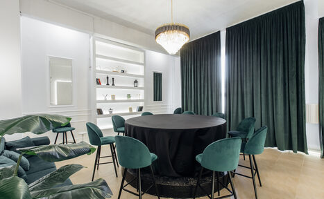 Арендовать Rubin Loft (Рубин Лофт). Зал Империал. Комната с круглым столом, покрытым черной скатертью, окруженным восемью зелеными стульями. На стене стеллаж с книгами и декором. Большие темные шторы, светлый пол. Центральная люстра. Зеленые растения в углу.