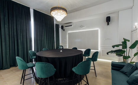 Арендовать Rubin Loft (Рубин Лофт). Зал Империал. Современное помещение с круглым столом, покрытым черной скатертью, девятью стульями с зелеными сиденьями, люстрой, зелеными шторами, кондиционером, светлым деревянным полом, декоративными элементами на стене, растением в горшке и нейтральным освещением.