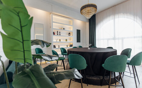 Арендовать Rubin Loft (Рубин Лофт). Зал Империал. Современное помещение с круглым столом, окружённым зелёными стульями. Наземные растения на переднем плане. На заднем плане встроенные полки с декором. Большое окно с прозрачными шторами. Подвесная люстра. Светлые стены и пол.