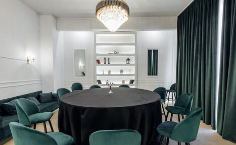 Арендовать Rubin Loft (Рубин Лофт). Зал Империал. Круглый стол с черной скатертью окружен бирюзовыми стульями. Справа зеленые шторы, слева диван. На стене встроенные полки с предметами, освещенные сверху. Центральное освещение — люстра. Цвета: белый, бирюзовый, зеленый, черный.