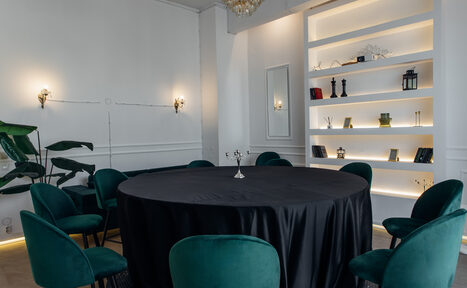 Арендовать Rubin Loft (Рубин Лофт). Зал Империал. Пространство с круглым столом, покрытым черной скатертью, окруженным зелеными стульями. На стене подсвеченные полки с декором, два бра и зеркало. Потолок с люстрой, белые стены, паркет. Зеленое растение сбоку.