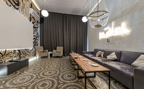 Арендовать Rubin Loft (Рубин Лофт). Зал Дубай. Просторная комната с диваном, креслами и длинным деревянным столом. На полу геометрический узор, стены с абстрактным рисунком и нейтрального цвета. Подвесные светильники и неоновая надпись на стене. Тёмные шторы закрывают окна.