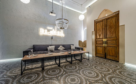 Арендовать Rubin Loft (Рубин Лофт). Зал Дубай. Лофт-пространство с серым диваном и журнальными столиками, серо-коричневые узорчатые плиточные полы, деревянные двустворчатые двери, декоративные светильники и неоновая надпись на стене.