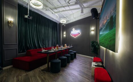 Арендовать Rubin Loft (Рубин Лофт). Лофт Вегас. Караоке-комната с темными стенами, длинный стол с черными пуфами, красные диваны, неоновая вывеска, шторы, подвесные светильники, большая картина с подсветкой, мягкое общее освещение, деревянный пол, декоративные растения.