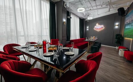 Арендовать Rubin Loft (Рубин Лофт). Лофт Вегас. Просторное помещение с большими окнами, затемнёнными шторами. Черный прямоугольный стол с красными стульями, сервирован посудой. Красный диван вдоль стены. Неоновая надпись на темной стене. Светлое деревянное напольное покрытие. Потолочные светильники и видимые коммуникации.