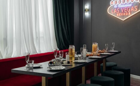 Арендовать Rubin Loft (Рубин Лофт). Лофт Вегас. Длинный обеденный стол с черной столешницей и сервировкой на шесть персон, черные пуфы вместо стульев. Красная скамья вдоль стены. Серый фон с неоновой надписью "Welcome to Fabulous Las Vegas Nevada". Белые и темные шторы. Настенные светильники.