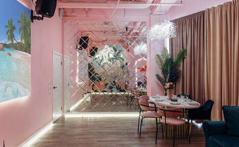 Арендовать Rubin Loft (Рубин Лофт). Лофт Пинки. Пространство с розовыми стенами, зеркальной стеной и настенным искусством с изображением пляжа. Деревянный пол, круглый стол и стулья, диван, декоративные растения, подвесные светильники. Потолок белый с открытыми коммуникациями. Фоновые шторы.