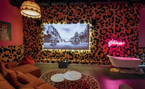 Арендовать Rubin Loft (Рубин Лофт). Лофт Гламур. Лофт с леопардовыми обоями, в центре большой экран, перед ним коричневый диван и круглый стол с мраморной столешницей. В углу белая ванна, на стене неоновая надпись "Glamour". Потолок черный, яркий абажур, полы плиточные.