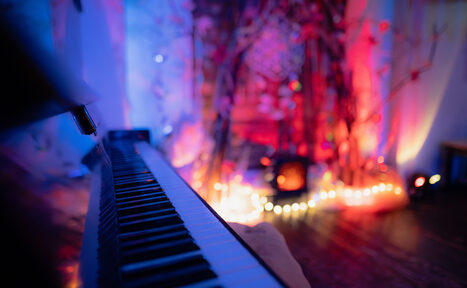 Арендовать Лес. Театральный лофт с верандой, сценой и вторым ярусом. Клавиши фортепиано слева на переднем плане. Позади него декоративная установка из веток и гирлянды с теплым освещением. Стены и пол темные, освещение разноцветное с синеватыми и красноватыми оттенками.