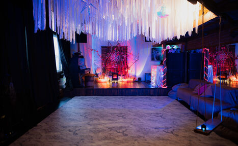 Арендовать Лес. Театральный лофт с верандой, сценой и вторым ярусом. Темное помещение с зоной отдыха. Конструкции и декор на потолке из светлых полос, мягкие диваны вдоль стены, яркая разноцветная подсветка. На возвышении декор с лампочками, стены кирпичные. Текстиль и элементы интерьера темных оттенков.