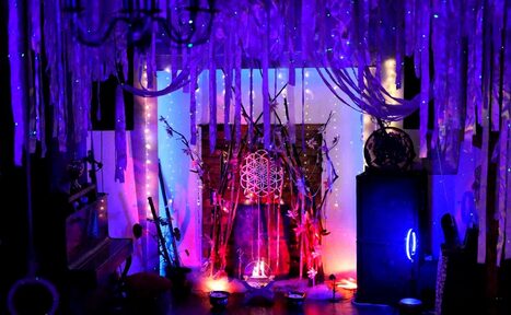 Арендовать Лес. Театральный лофт с верандой, сценой и вторым ярусом. Темное пространство с яркой подсветкой; гирлянды светодиодов в фиолетовых и голубых тонах. На полу низкие мисочки, по центру узор внутри деревянной конструкции. Потолок декорирован свисающими тканями, справа круглое зеркало, слева пианино.