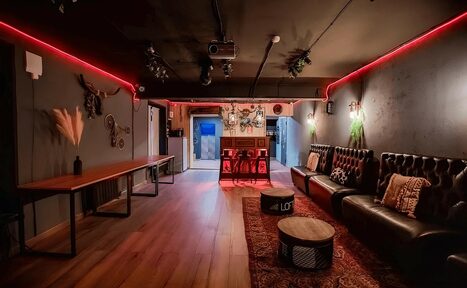 Арендовать Пространство для любых мероприятий с верандой и грилем. Лофт-пространство с деревянным полом, кожаным диваном, деревянными столами. Красная подсветка на стенах, потолок с прожекторами. На стенах декор, подушки на диване. Барная стойка с напитками, два круглых стола. Тёмные стены.