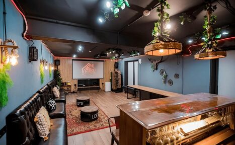 Арендовать Пространство для любых мероприятий с верандой и грилем. Лофт-пространство с барной стойкой, длинным столом, диванами, пуфами, проектором, экранами. Потолочные светильники, декоративные элементы и растения. Деревянный пол, стены синие, потолок темный.