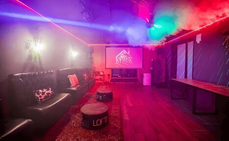 Арендовать Пространство для любых мероприятий с верандой и грилем. Лофт-пространство с черными кожаными диванами, маленькими столиками, большим экраном и надписью "LOFT". Темное освещение с цветными световыми эффектами, дым. Деревянный пол и стол, ковёр. Светильники на стенах.