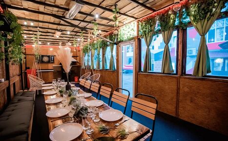 Арендовать Пространство для любых мероприятий с верандой и грилем. Помещение с деревянными стенами и потолком, продолговатый стол с сервировкой для банкета на много гостей. Металлические и деревянные стулья, зелёные шторы и декор из растений. Настенные светильники, дартс на стене, подвесные кресла и диван вдоль стены.