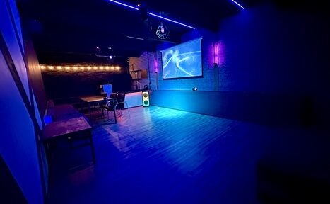 Арендовать Kahovka loft (Каховка Лофт). Неоновый лофт-зал. Комната с темными стенами, деревянным полом, столами и стульями слева. Напротив висит экран с проектором. На стенах неоновое освещение и светящиеся декоративные элементы. В углу стоит колонка.