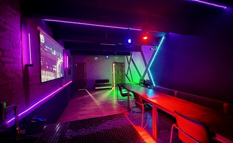 Арендовать Kahovka loft (Каховка Лофт). Неоновый лофт-зал. Темное помещение с неоновыми акцентами. Справа длинный деревянный стол с разноцветными стульями. На стенах и потолке неоновые ленты фиолетового и зеленого цветов. Экран на левой стене, деревянный пол и двери.